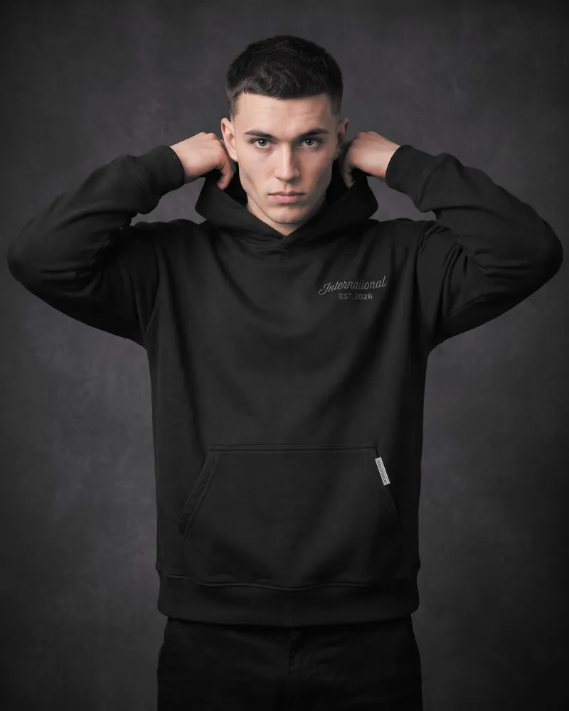 A young man with a short dark crop, both hands gripping the hood at his neck, wearing the black International hoodie, dark grey International script on the chest and a silver bar on the pocket