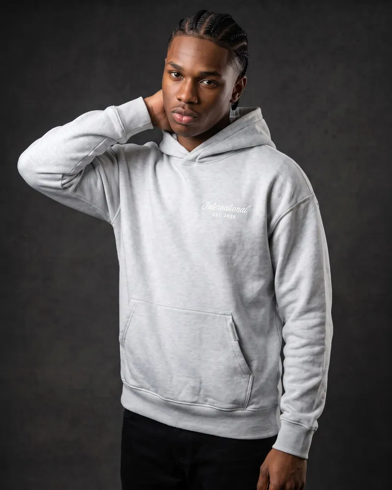 A young man with fresh cornrow braids, one hand behind his neck, wearing the ice grey International hoodie with white International script on the chest and a silver bar on the pocket