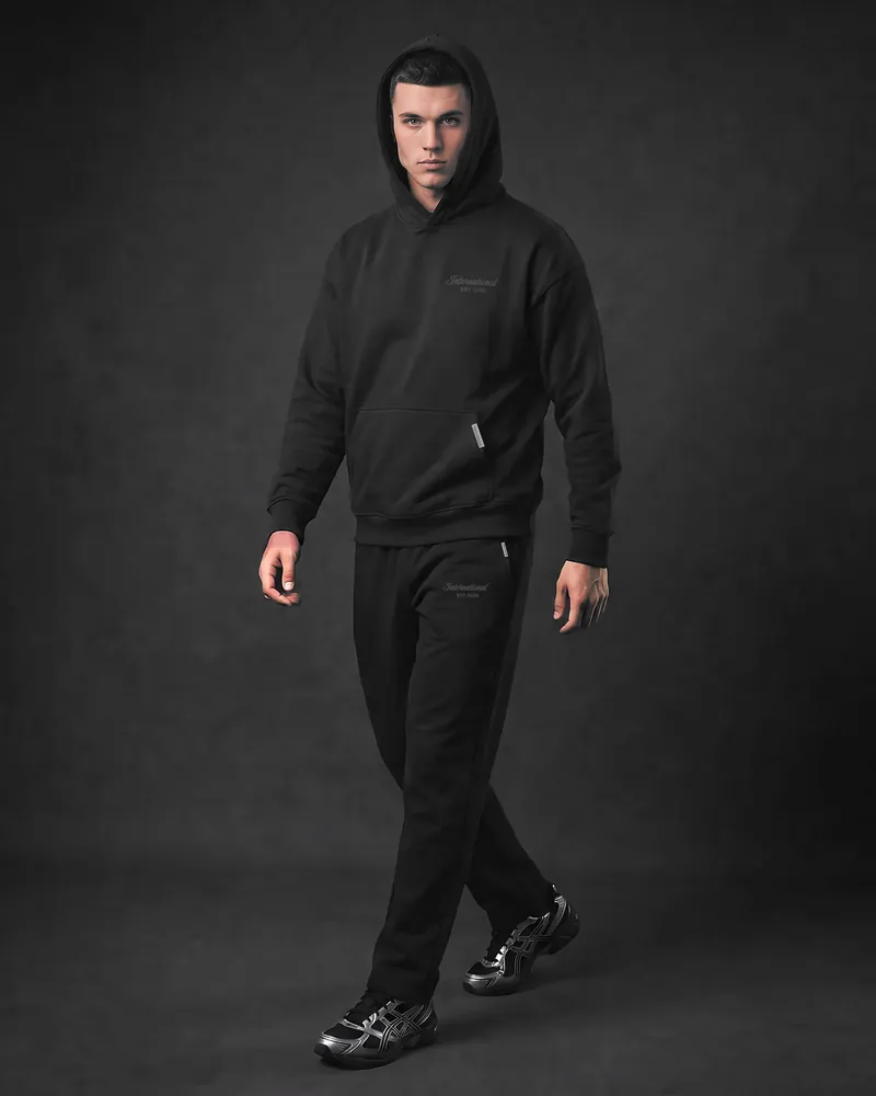 A young man walking towards the camera with his hood up, in the black International hoodie and matching black International joggers, the International script on the left thigh, a silver bar on the pocket and black and silver trainers