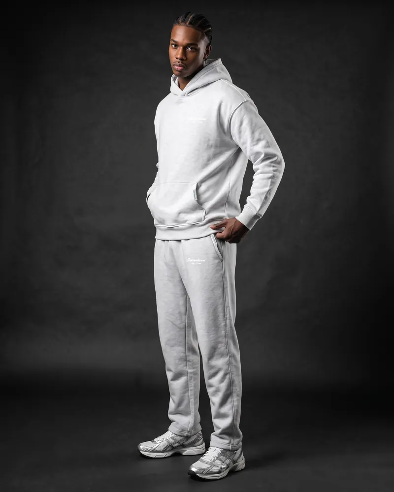 A young man with fresh cornrow braids standing three-quarter on, hand in the hoodie pocket, in the ice grey International hoodie and matching ice grey joggers, white International script on the left thigh, a silver bar on the pocket and white and silver trainers