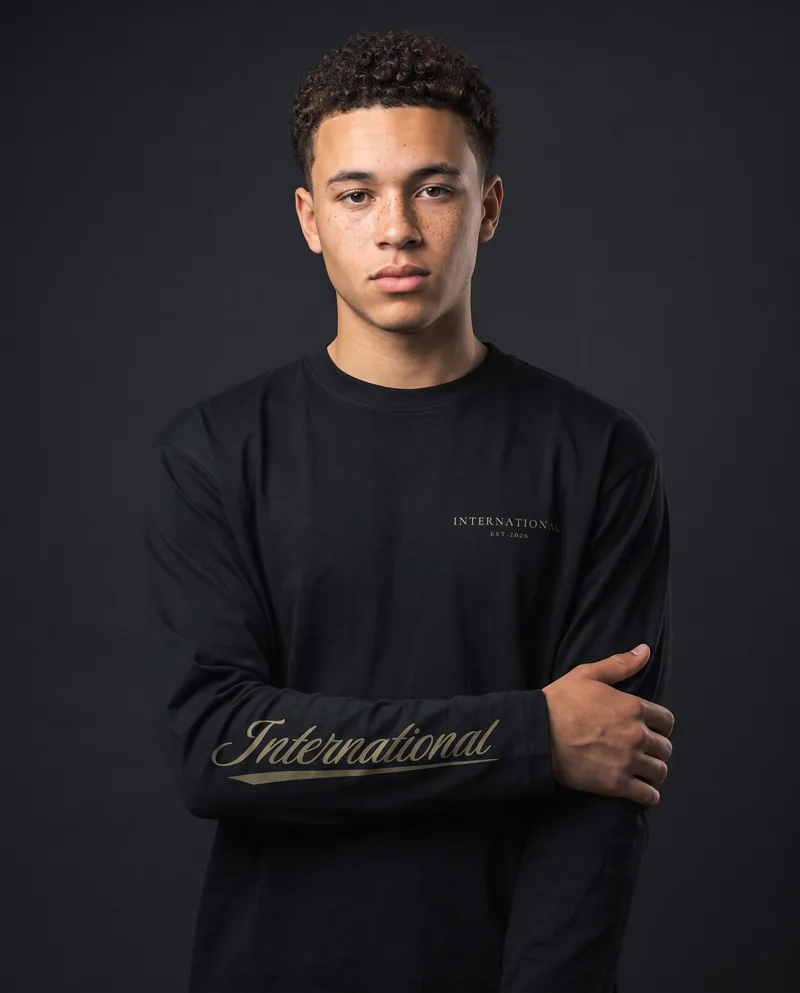 A young man with short curly hair and freckles in the black International long sleeve, khaki INTERNATIONAL EST.2026 on the chest and the khaki International script along his right forearm