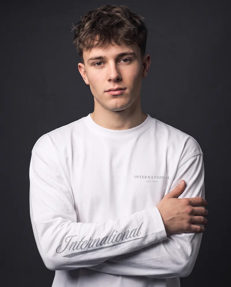 A young man with a messy brown fringe, arms crossed, in the white International long sleeve, silver INTERNATIONAL EST.2026 on the chest and the silver International script along his right forearm